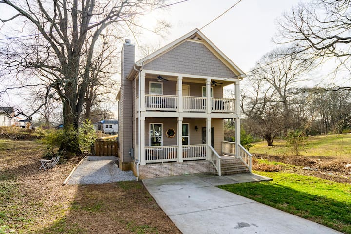 Two-story house with a beige exterior, white railings on two front porches, a driveway, and surrounded by trees, located in a suburban setting under bright daylight.