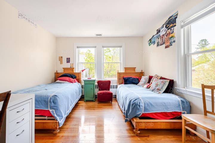 Bright bedroom with two twin beds dressed in blue and red linens, a red armchair, and a green nightstand centered between the beds. Sunlight streams through large windows with tree views, creating a warm and inviting atmosphere.