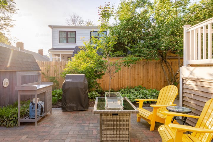 A backyard patio with two yellow Adirondack chairs, a grill, and a fire pit table surrounded by greenery, ideal for hosting a tailgate gathering.