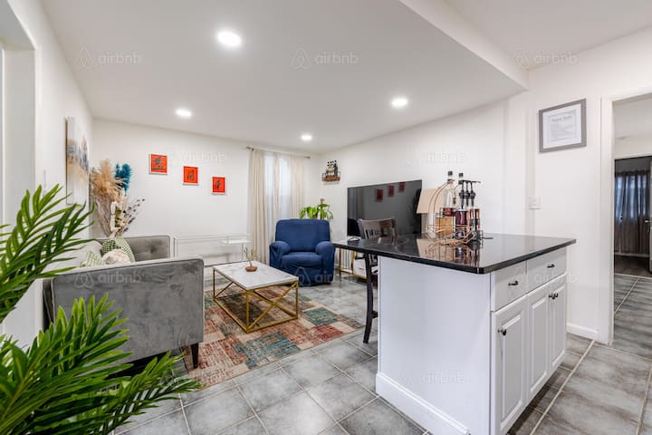 Bright and modern living space with a kitchen island, a cozy seating area featuring a blue armchair and gray couch, and colorful wall art, perfect for relaxing before or after a football game.