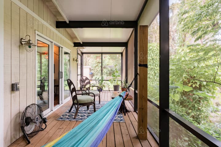 A cozy screened porch with wooden floors featuring a colorful hammock, wicker chairs, a small table, and lush greenery outside, perfect for relaxing after a tailgate in Atlanta.
