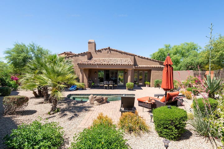 A backyard of a vacation rental featuring a small pool surrounded by desert landscaping, lounge chairs, and a red umbrella under a clear blue sky.