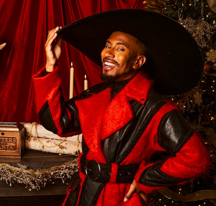 A person dressed in a festive red and black cowboy-style outfit with a wide-brimmed black hat, standing in front of a holiday-themed backdrop featuring a decorated Christmas tree, candles, and a box labeled 'Letters to Santa.'