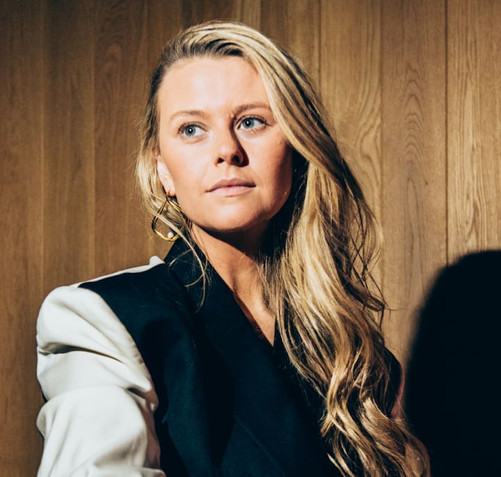 A person with long wavy blonde hair wearing a black and white jacket, standing against a textured wooden wall in warm lighting.