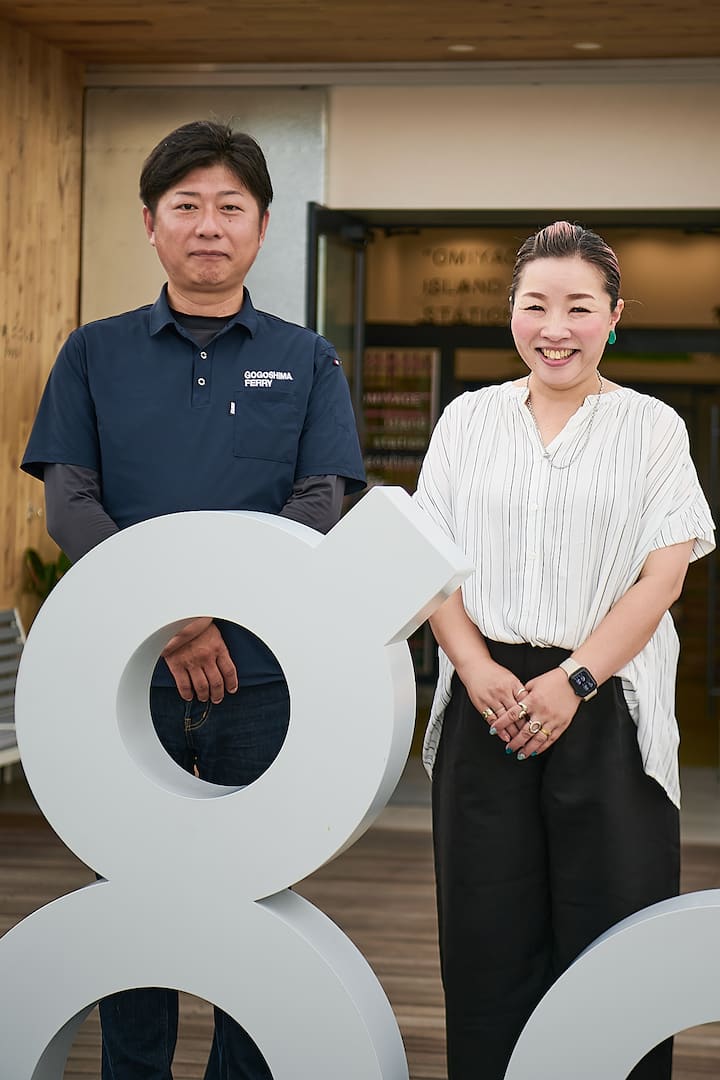 Two people standing behind a white sculpture shaped like the number eight in front of a building entrance, with 'Gogo Shima Ferry' written on one person's shirt.