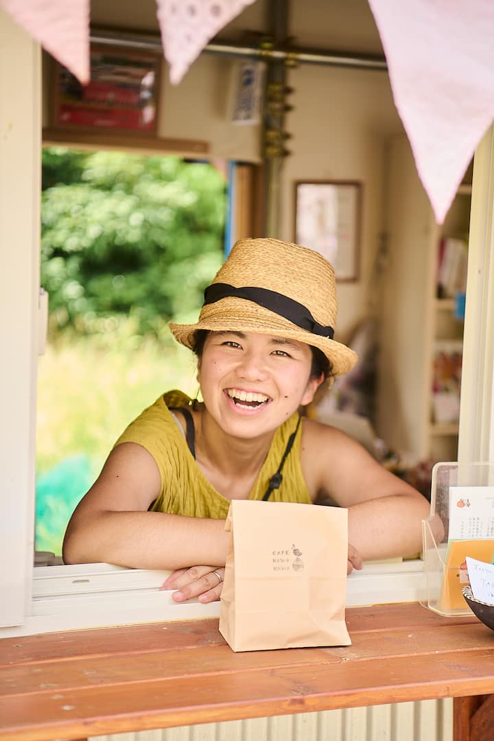Close-up of a cafe window counter featuring a paper bag with 'Cafe Nan Nan' logo, surrounded by light pink bunting decorations and a blurred green outdoor background.