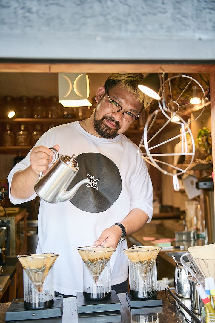 A person pours hot water into coffee drippers at a small, cozy café on a Japanese island, surrounded by jars and warm lighting.