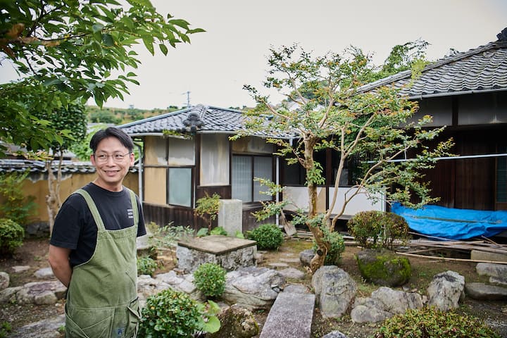 A traditional Japanese home with a tiled roof surrounded by a lush garden, featuring rocks, shrubs, and a small tree, located on Gogoshima Island.