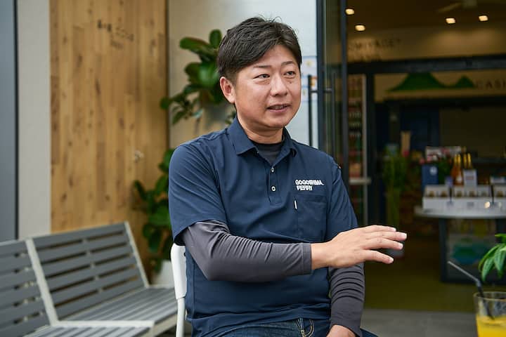 A person wearing a navy blue polo shirt with 'Kagoshima Ferry' embroidered on it, seated outdoors near a wooden wall and greenery, gesturing with their hand.