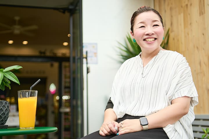 A person seated at an outdoor café table, wearing a white striped blouse and a smartwatch, with a glass of orange juice and a potted plant on the table. A blurred background shows the entrance to a store with warm lighting and wooden decor.