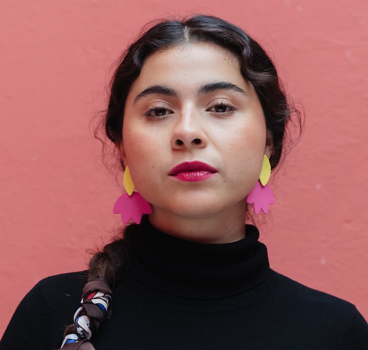 A close-up of a person wearing a black turtleneck sweater, colorful earrings, and a braided hairstyle with a vibrant pink wall in the background, located in Mexico City.