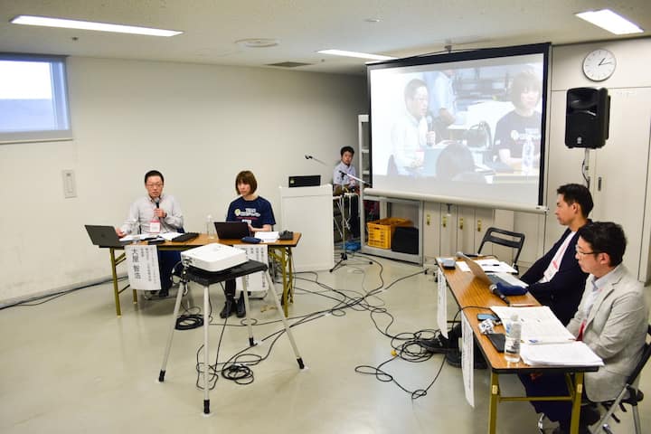 A small conference room with two tables, a projector, and a screen displaying a panel discussion. Cables run across the floor, and participants are seated, engaging in a discussion.