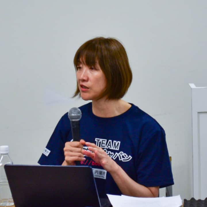 A person wearing a dark blue shirt with 'TEAM 防災ジャパン' printed on it, holding a microphone and sitting in front of a laptop and papers on a desk, giving a presentation.