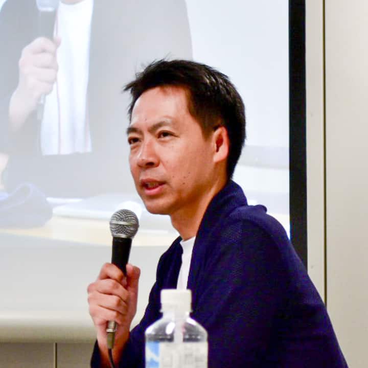 A person holding a microphone during a presentation, with a projection screen visible in the background showing the same person speaking.