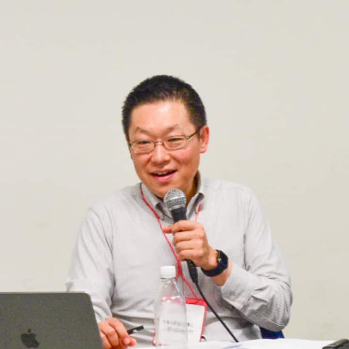 A speaker wearing a light-colored shirt and red lanyard holds a microphone, seated at a table with a laptop and a bottle of water during a discussion about disaster preparedness and alternative evacuation solutions.