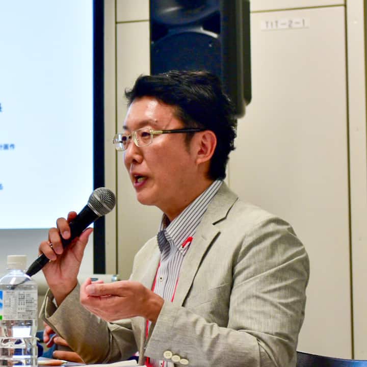 A person in a gray suit sits at a desk holding a microphone and gesturing during a presentation, with a large speaker and projector screen visible in the background.