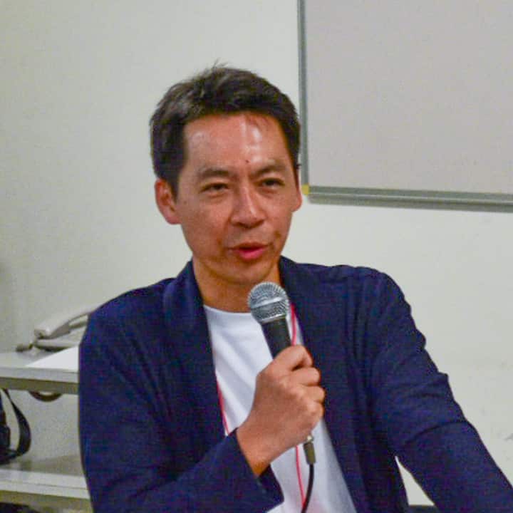 A speaker in a navy jacket holding a microphone, standing in front of a whiteboard during a presentation about disaster preparedness and support systems at an event.