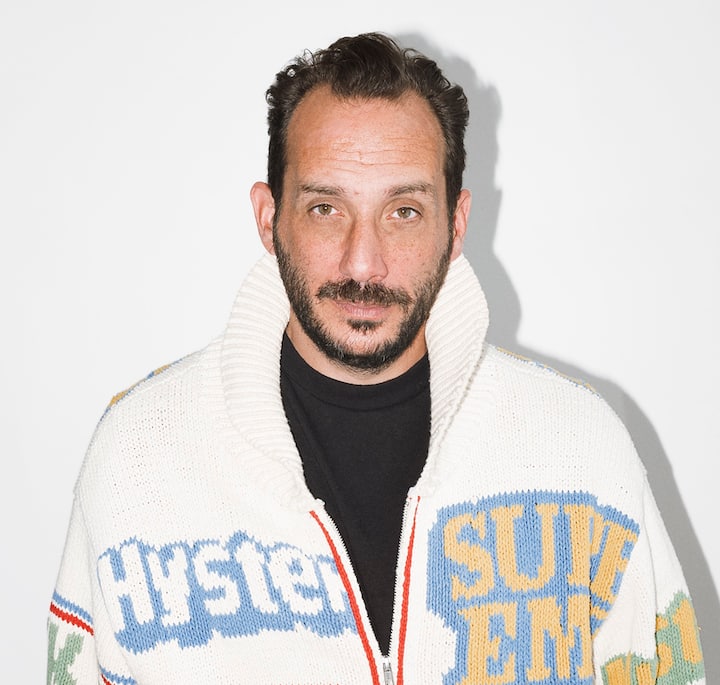 A person wearing a white sweater with colorful text patterns, including the words 'Hysteric' and 'Supreme' in bold font, standing against a plain white background.