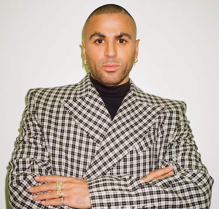 A person wearing a black turtleneck and a checkered blazer with arms crossed, showcasing bold gold rings on their fingers against a plain white background.