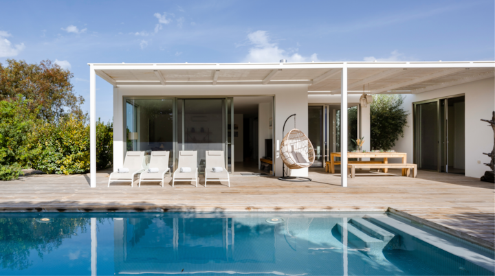 Maison de vacances moderne avec une piscine, une terrasse en bois, trois chaises longues, un fauteuil suspendu et un coin repas extérieur entouré de verdure sous un ciel bleu sans nuages.