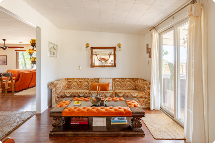 Un salon lumineux à la décoration rétro avec un canapé à motifs orange et marron, et une table basse en bois avec matelassage orange et plateau en verre. Un miroir est suspendu au centre du mur blanc, et la lumière du soleil traverse les baies vitrées coulissantes sur la droite.
