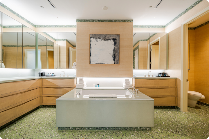 Salle de bain moderne avec une grande baignoire îlot au centre, entourée d'éléments en bois, de miroirs et d'un sol en carrelage vert. Des serviettes et des articles de toilette sont soigneusement disposés sur les bords de la baignoire.