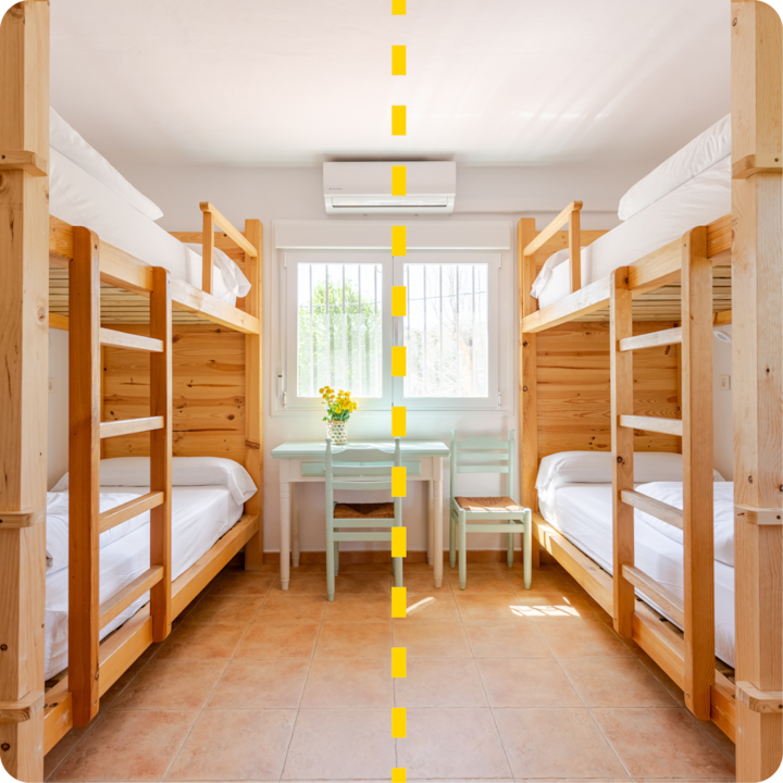 Pièce lumineuse avec deux lits superposés en bois de chaque côté, séparés par une ligne pointillée jaune, avec une petite table, un vase de fleurs jaunes et des chaises placées devant une fenêtre au centre.