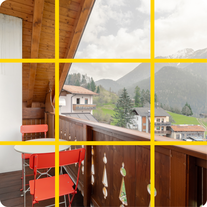 Un balcon confortable avec des chaises rouges et une petite table, surplombant un village alpin pittoresque avec des chalets, une végétation luxuriante et des montagnes au sommet enneigé au loin.