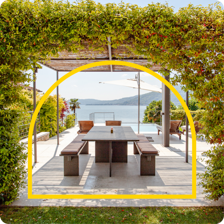 Un coin repas extérieur à l'ombre d'une pergola en bois couverte de verdure, surplombant un lac paisible et des collines en arrière-plan, encadré par une arche jaune.