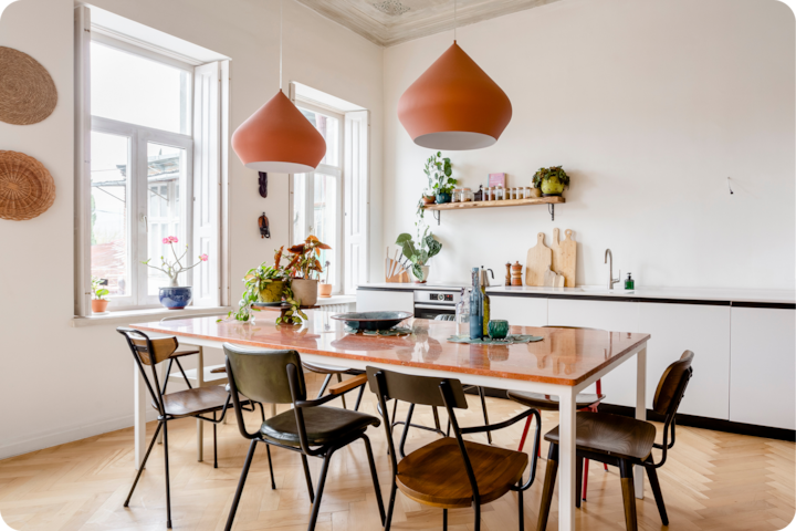 Cuisine lumineuse avec une grande table en bois entourée de chaises dépareillées, des suspensions orange et des plantes sur la table et les étagères.
