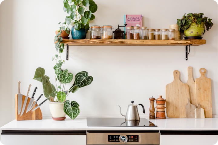 Une cuisine minimaliste avec une étagère en bois sur laquelle sont posés des bocaux, des plantes et un livre de cuisine rose, au-dessus d'un plan de travail où se trouvent une plante, des couteaux, une bouilloire et des planches à découper.