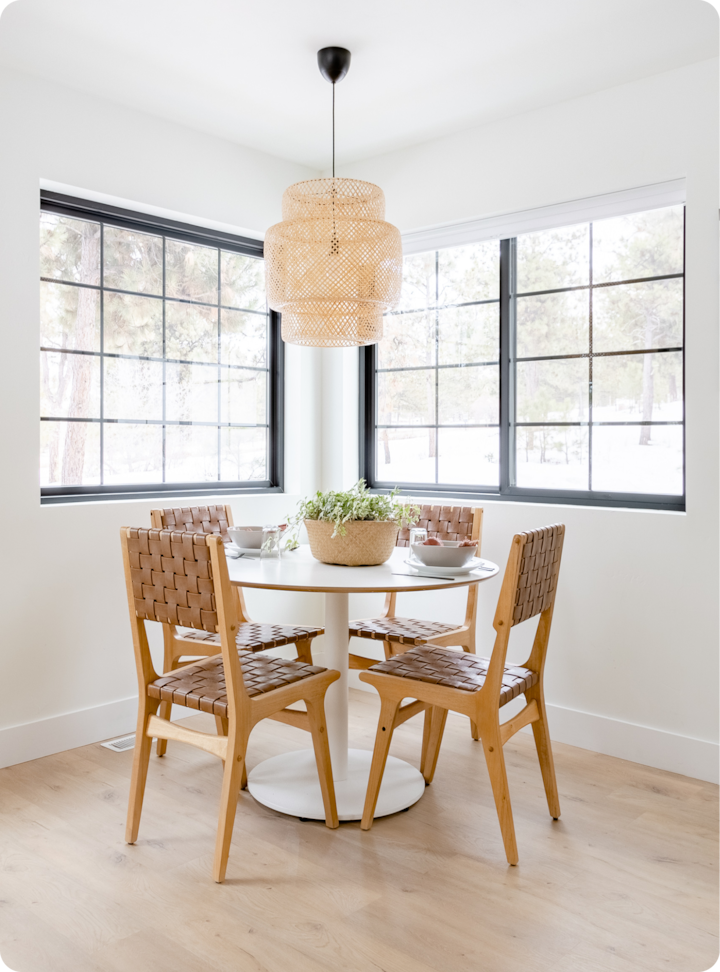 Un coin repas confortable avec une table ronde blanche, quatre chaises en bois tressé et une plante en pot au centre, entouré de grandes fenêtres donnant sur des arbres enneigés.