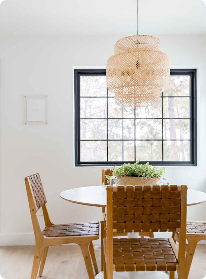 Un coin repas confortable avec des chaises en bois tressé, une table ronde blanche et un panier de plantes vertes sous une suspension en osier. Une fenêtre aux encadrements noirs donnant sur les arbres en arrière-plan.
