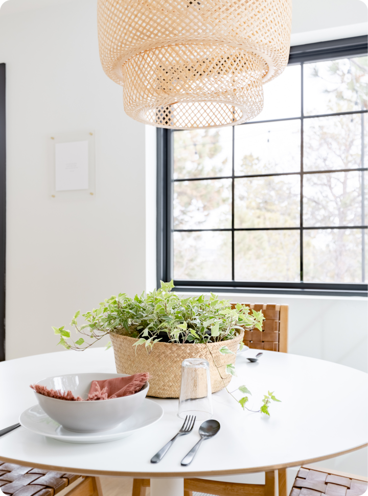Une salle à manger lumineuse avec une table ronde blanche, un panier tressé de plantes vertes, un bol soigneusement dressé avec une serviette terracotta et des couverts noirs placés à côté d'un verre, sous un luminaire en rotin tissé naturel avec une fenêtre moderne en arrière-plan.