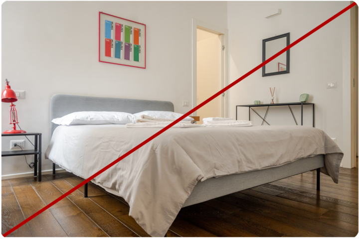 Chambre minimaliste avec un lit soigneusement fait, une tête de lit grise, des draps blancs et des serviettes pliées posées dessus. Une lampe de chevet rouge et des tableaux ajoutent des touches de couleur à la vaste pièce lumineuse. La hauteur de l'appareil met en valeur l'agencement de la pièce, y compris le parquet et une petite console contre le mur du fond.