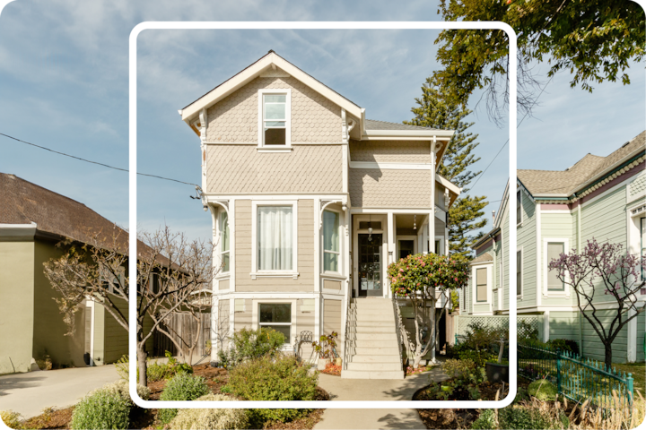 Vue extérieure d'une maison de deux étages gris clair avec un jardin paysager par une journée ensoleillée, entourée d'autres maisons.