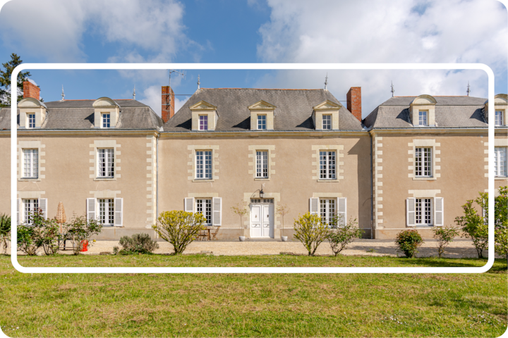 Un bâtiment champêtre beige symétrique de style français avec un toit en ardoise, entouré d'arbustes taillés et d'une pelouse vert vif sous un ciel partiellement nuageux.