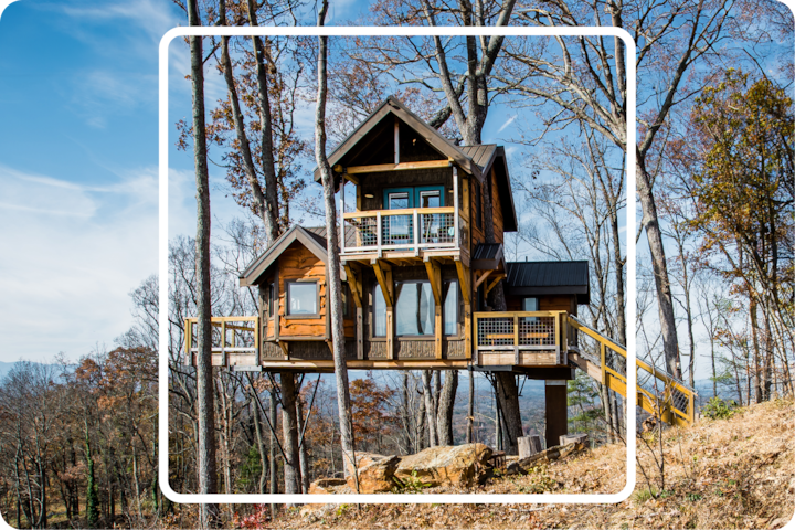 Cabane perchée rustique à plusieurs niveaux avec de grandes fenêtres et des balcons en bois, nichée parmi de grands arbres dans une forêt, avec vue sur des montagnes au loin par une journée ensoleillée.