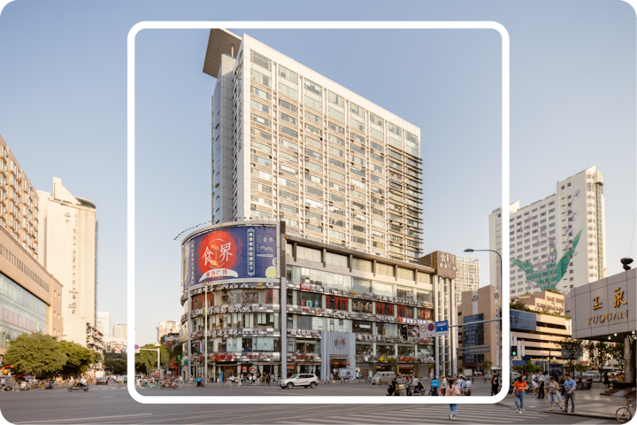 Carrefour urbain animé avec des piétons et des véhicules, dominé par un immeuble moderne à angle arrondi et un grand panneau publicitaire affichant des caractères chinois, sous un ciel bleu sans nuages.