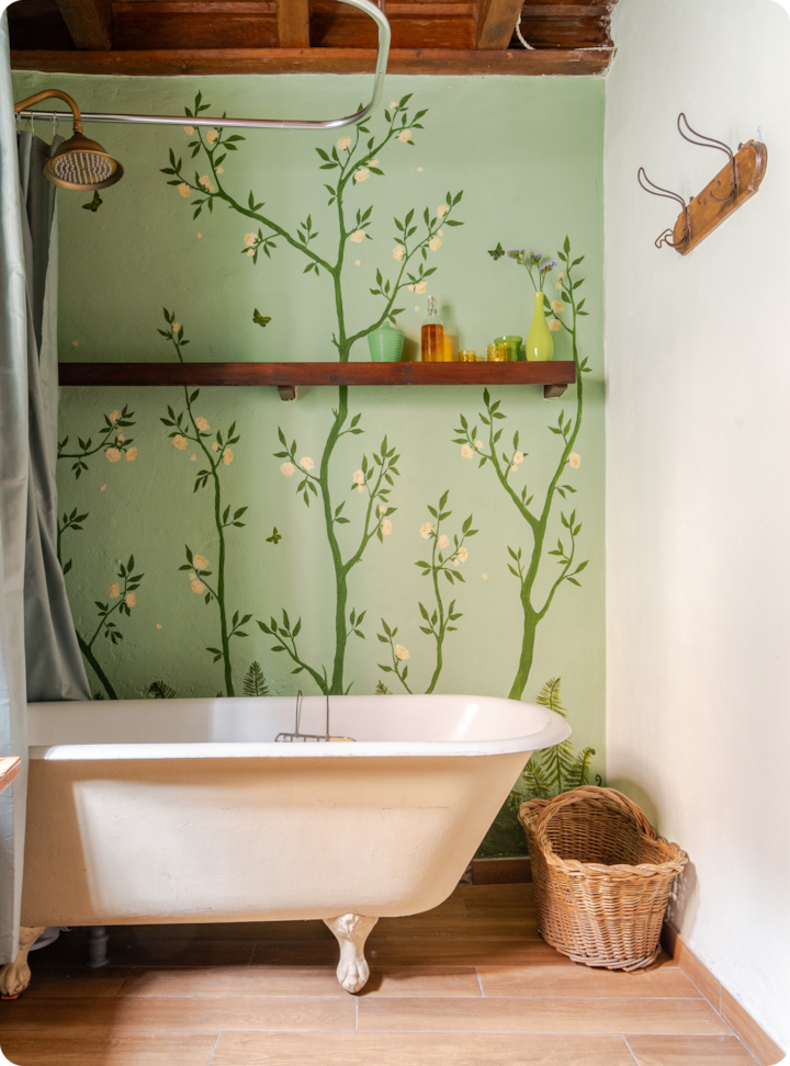 Une salle de bain confortable avec une baignoire sur pieds, un mur vert peint avec un motif floral, une étagère en bois contenant des bouteilles en verre colorées et un panier en osier sur le parquet.