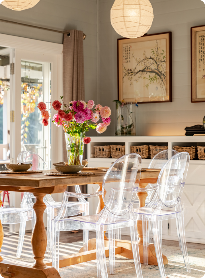 Salle à manger baignée d'une lumière douce meublée d'une table en bois dressée avec des sets, entourée de chaises en acrylique transparent. Un bouquet de fleurs coloré dans un vase en verre est placé au centre de la table. En arrière-plan, des paniers en osier sont soigneusement rangés sur des étagères et des tableaux de style japonais ornent le mur.