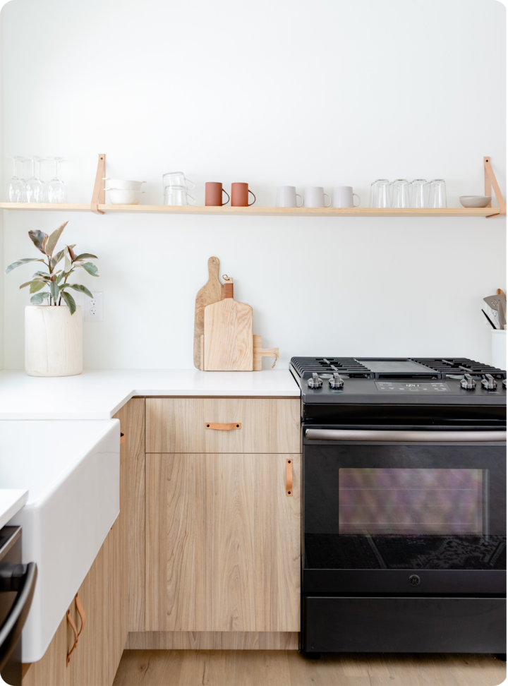 Coin cuisine moderne avec des placards en bois clair, une cuisinière à gaz noire, un évier de ferme blanc et des étagères ouvertes minimalistes contenant des mugs, des verres et des objets de décoration.