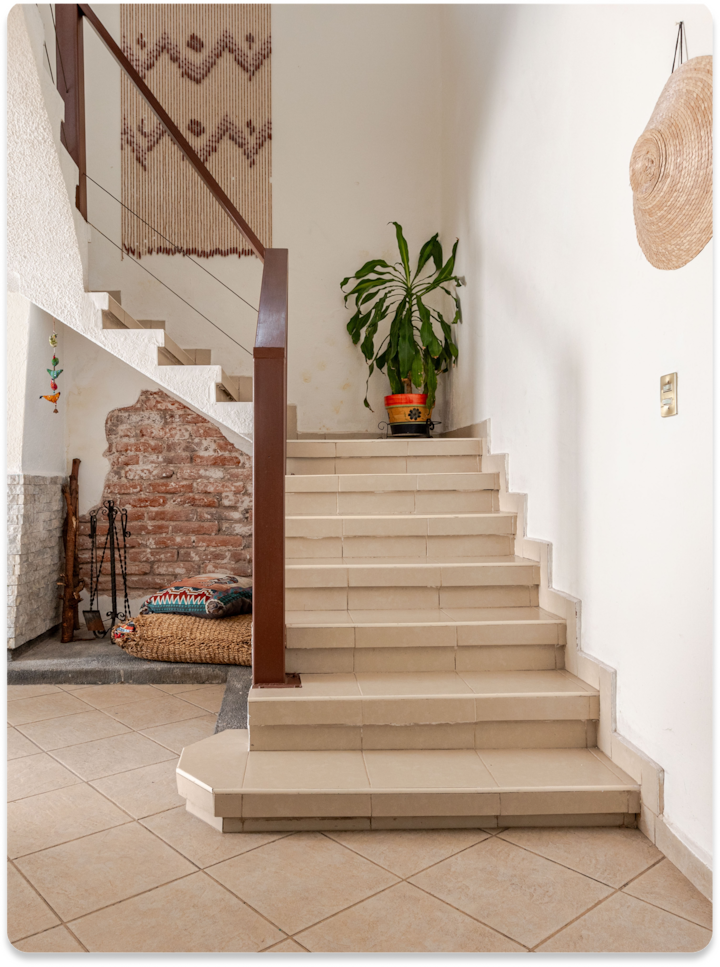 Escalier intimiste en carreaux beiges, plante verte en pot sur le palier, tenture murale tissée avec des motifs géométriques et mur de briques rustique sur la gauche.