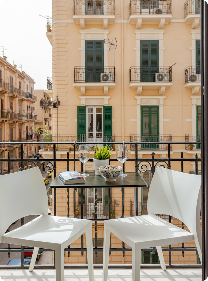Balcon confortable avec une petite table pour deux, des chaises blanches, des verres à vin, une plante en pot et un livre, donnant sur un immeuble de style européen ensoleillé avec des volets verts.