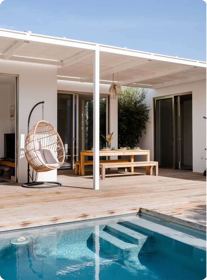 Terrasse avec une chaise en osier suspendue, une table en bois et une piscine à l'eau transparente sous un ciel bleu.