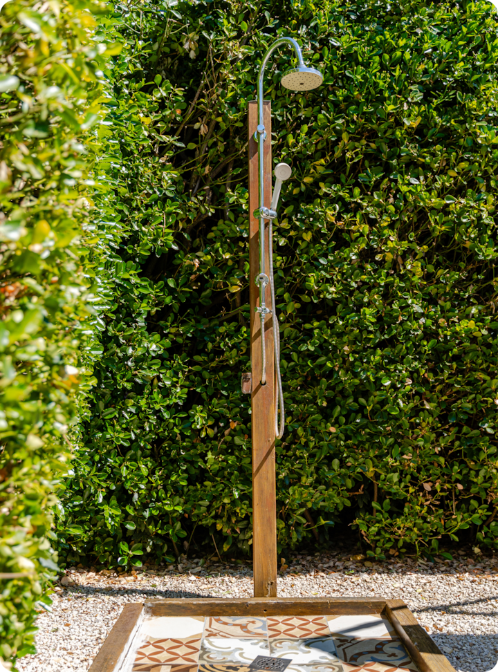Douche extérieure entourée de haies verdoyantes, avec un poteau en bois surmonté d'une pomme de douche et des carreaux aux motifs travaillés en dessous.