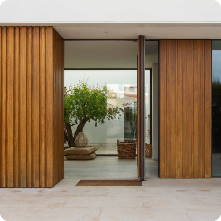 Entrée principale accessible avec une large porte en bois et un sol en pierre plate sans marches menant à un logement moderne. À l'intérieur, on peut voir un arbre en pot et des paniers en osier sur un sol lisse.