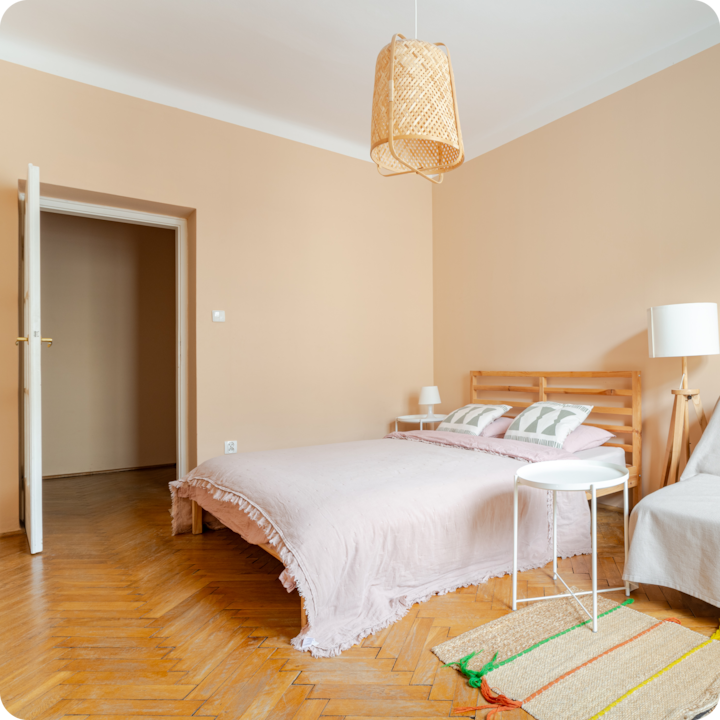 Chambre confortable avec un lit en bois, une literie rose pastel, une table d'appoint ronde blanche et un mur beige. Le sol présente un motif à chevrons et une porte ouverte révèle une pièce adjacente.