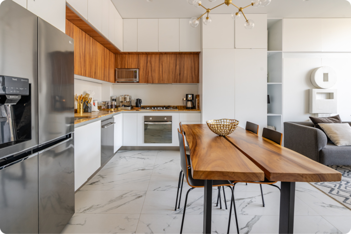 Cuisine et salle à manger modernes avec un réfrigérateur en acier inoxydable, des éléments en bois, un sol en marbre, une table en bois entourée de chaises noires et un salon confortable en arrière-plan.