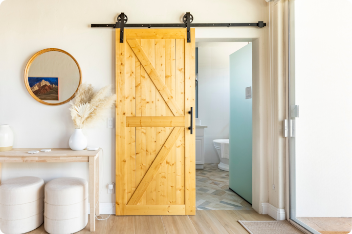 Une pièce confortable avec une porte coulissante en bois menant à une salle de bain, une console en bois clair, un miroir rond et des herbes de la pampa sur la gauche.